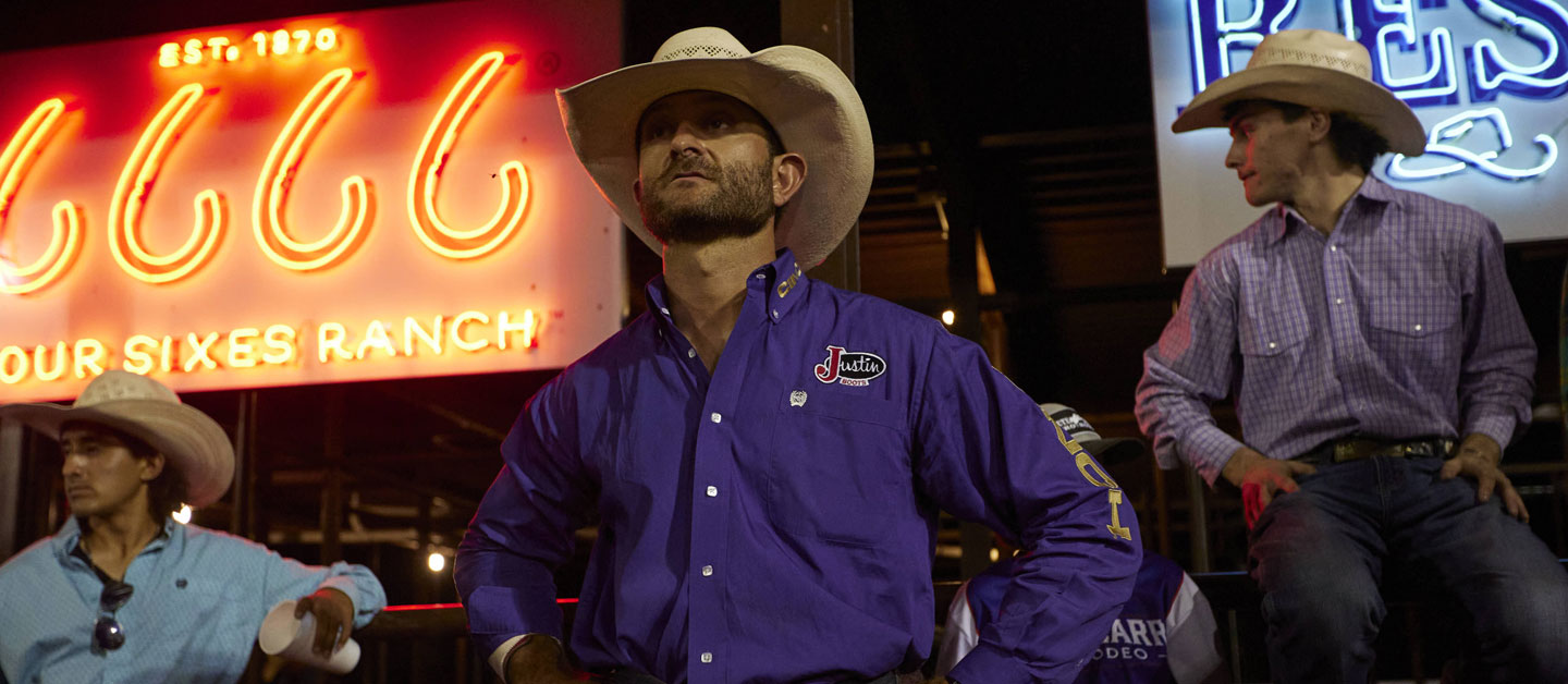 Man wearing a purple shirt and a cowboy hat standing in front of a big screen at a rodeo.